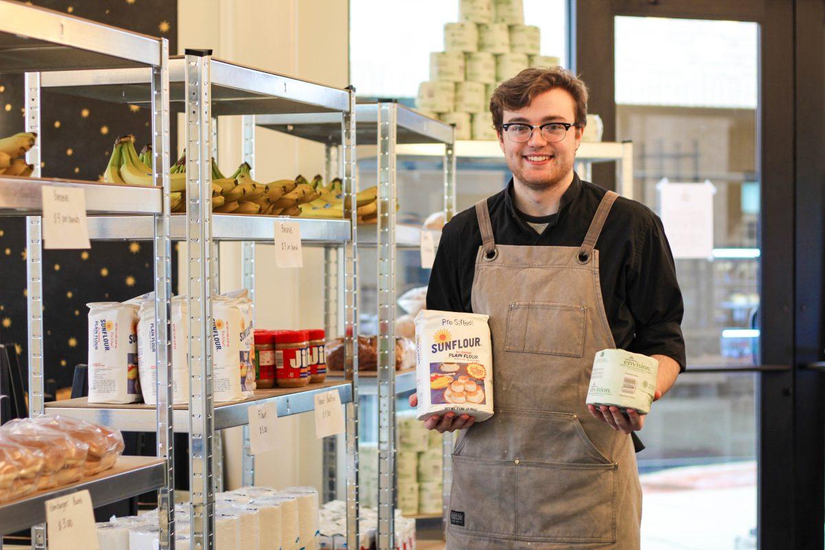 James May, barista at Strange Brew Coffeehouse, holds toilet paper and flour, two of the necessity items the business is selling to the public.